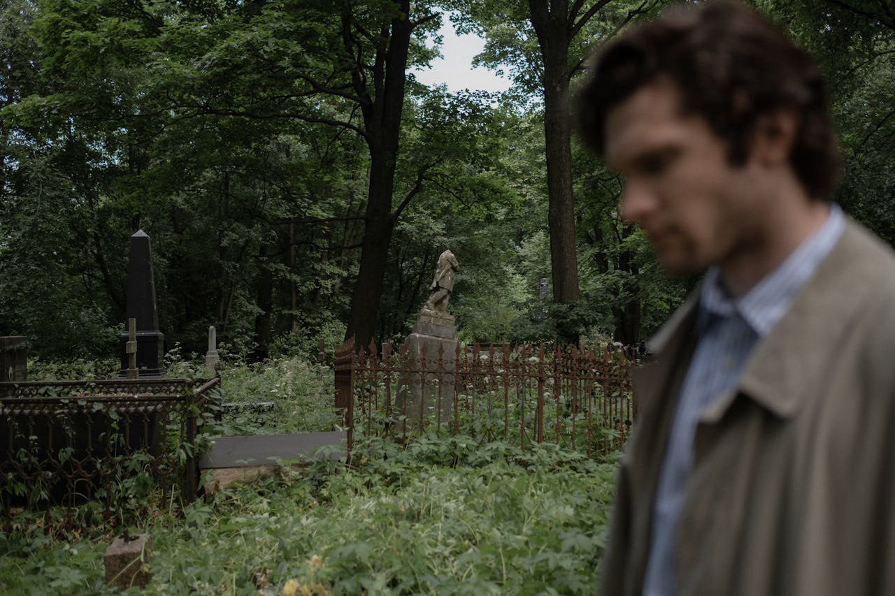 A blurred man in a trench coat stands near old gravestones in a lush summer cemetery setting.