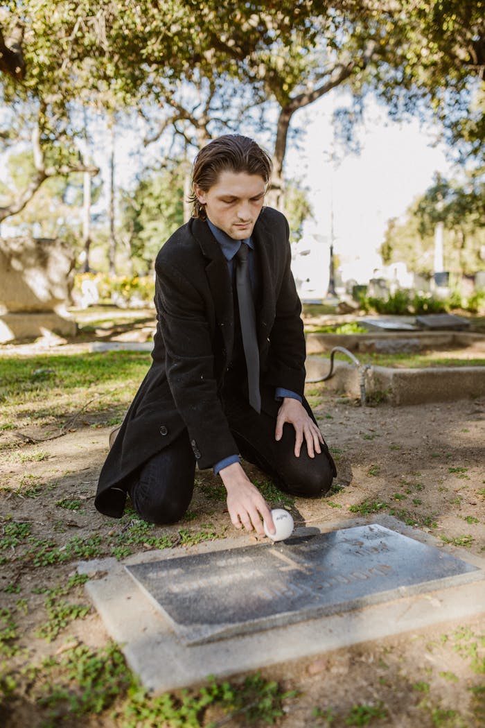A man in a black coat kneeling at a gravestone in a cemetery during the day, paying respects.