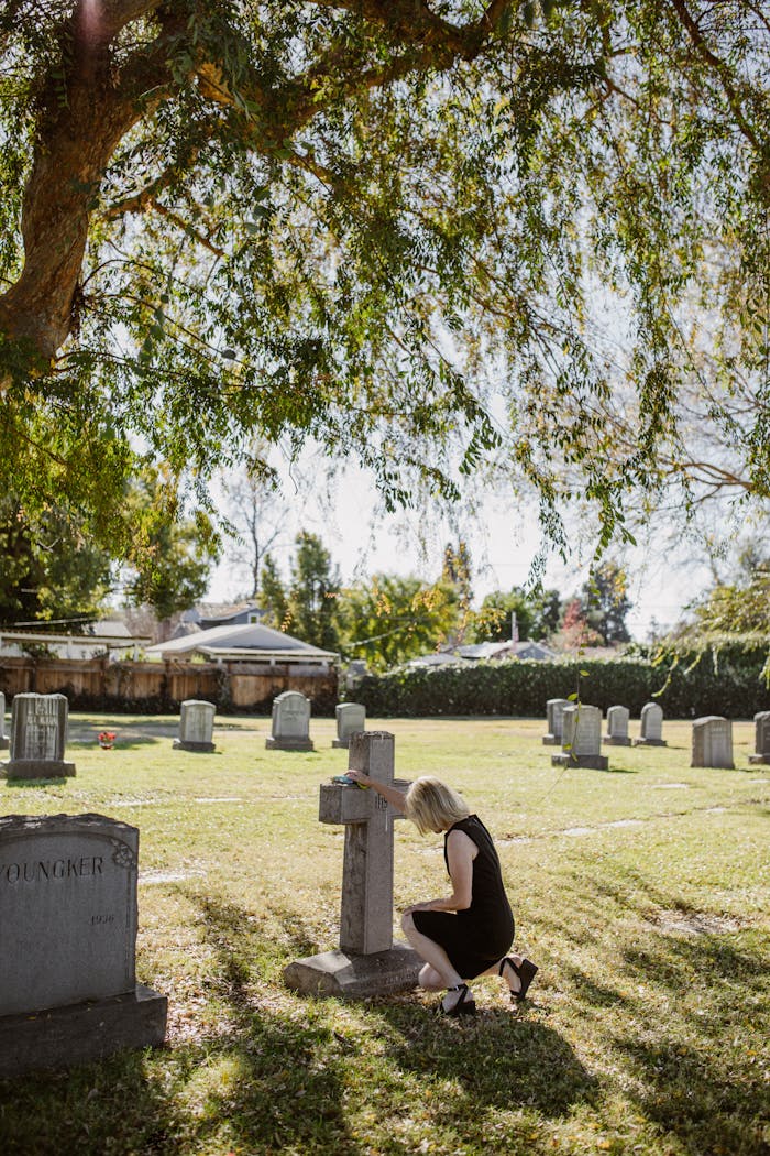 A somber scene of a woman kneeling by a gravestone in a peaceful cemetery during a sunny autumn day.