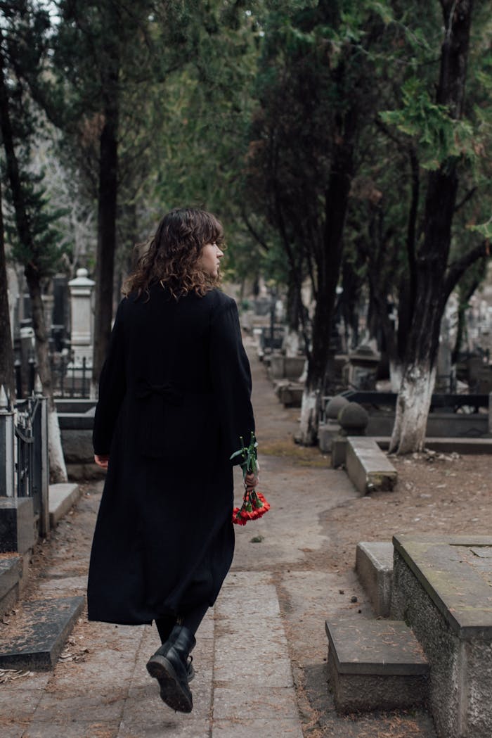 A person in black attire holding flowers, walking along a cemetery pathway surrounded by trees.
