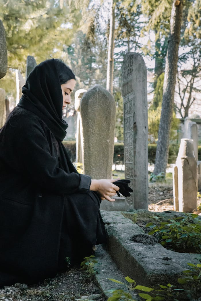 Side view full body of calm ethnic female with gloves wearing black clothes and headdress squatting near shabby tombstone on cemetery