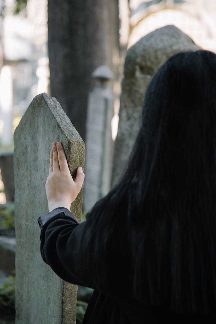 portfolio-07 Back view of faceless female in black clothes touching old stone gravestone while spending time on cemetery on blurred background